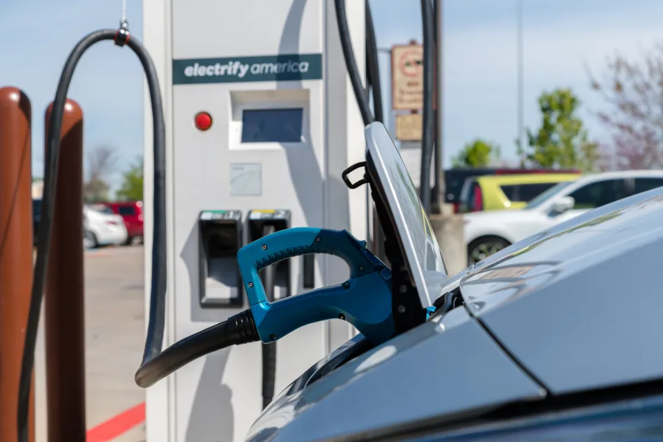 An electric vehicle is plugged into a charging station. A sticker reads “charge America” on the station.